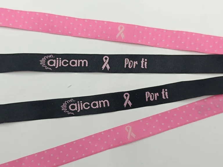 Pulsera tejida personalizada en textil rosa y negro con logotipo de AJICAM y lazo contra el cáncer de mama, fabricada en tejido resistente.